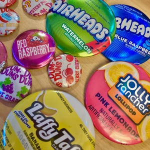 May include: A collection of colorful candy-themed buttons. The buttons feature various flavors like watermelon, blue raspberry, pink lemonade, and banana. Some buttons have the brand names Airheads, Jolly Rancher, and Laffy Taffy.