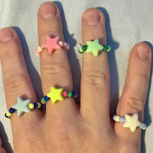 May include: A hand displays four beaded rings, each featuring a star-shaped charm. The rings showcase a variety of colors, including pink, green, yellow, and blue, with the beads complementing the star colors. The rings are a fun and colorful accessory.