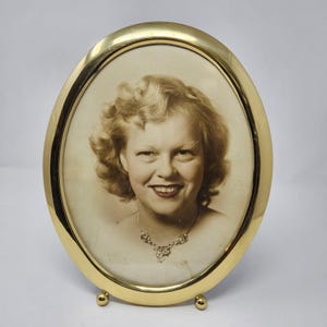 May include: An oval-shaped, polished brass picture frame featuring a smooth, reflective metallic border. The frame contains a vintage-style, sepia-toned portrait photograph of a smiling person with wavy blonde hair wearing a delicate necklace.