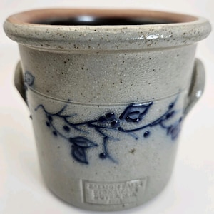 May include: A stoneware crock with a light gray base and a dark brown rim. The crock features a blue floral design and two small handles. The text "Salmon Falls Stoneware Dover NH" is embossed on the front.