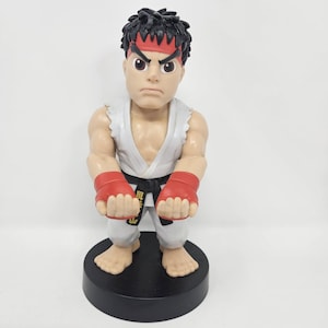 May include: A stylized figurine of a martial artist with spiky black hair and a red headband. The figure wears a white karate gi with a black belt featuring gold Japanese characters, red hand wraps, and stands on a circular black base.