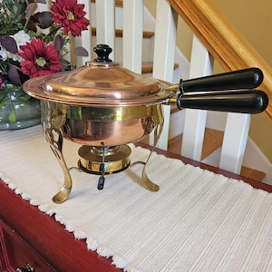 May include: A copper and brass chafing dish with a black handle and lid knob. The dish has a round base with a burner and decorative legs. The lid is copper, and the body is brass. The handles are black.