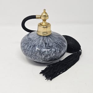 May include: A round glass perfume bottle featuring a black and white mottled pattern. The bottle is topped with a gold-toned metal atomizer pump, which is connected to a black fabric-covered bulb and a long black tassel hanging from the side.