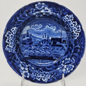 May include: A blue and white ceramic plate featuring a transferware scene of ships and a fort, with the text "LANDING OF GEN LA FAYETTE AT CASTLE GARDEN NEW YORK 16 AUGUST 1824" printed at the bottom. The border is decorated with floral patterns.