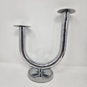 May include: A U-shaped chrome metal candle holder featuring a polished reflective surface. The object has a circular base with concentric ring detailing and two vertical arms, each topped with a flat circular plate for holding candles.