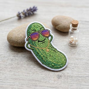 May include: Embroidered patch featuring a cool pickle wearing sunglasses and a gold chain with a star pendant. The pickle is green with a white border. The sunglasses have purple and orange lenses. A fun, quirky accessory.