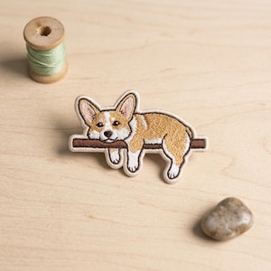 May include: Embroidered patch featuring a corgi dog lying on a brown log. The corgi has tan and white fur with a detailed stitched design. A wooden spool of green thread and a small stone are also in the image.