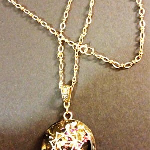 May include: Gold-toned necklace with a circular pendant. The pendant features a black background with gold and purple accents and a white geometric design. The chain is a delicate link style. A decorative bail connects the pendant to the chain.