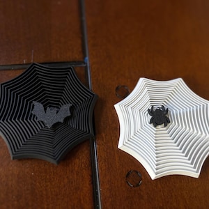 May include: Two hexagonal coasters featuring a raised spiderweb pattern. One coaster is solid black with a central black bat silhouette, while the other is solid white with a central black spider figure. Both items have a layered, geometric design.