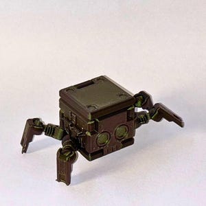 May include: A small, dark olive green robotic creature with a cube-shaped body and four articulated legs. The front of the cube features two circular eye-like indentations and a textured surface, with a flat top and mechanical joints.