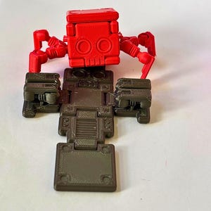 May include: A red articulated robot figure with a blocky head and multiple jointed legs, positioned above a dark brown, flat, rectangular base with segmented panels and mechanical details. The figure features a skull-like face design.