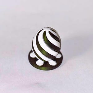 May include: A small, rounded decorative object featuring a swirling pattern of alternating dark olive green and white stripes. The object is mounted on a circular, flat dark olive green base with small white accents visible at the bottom edge.