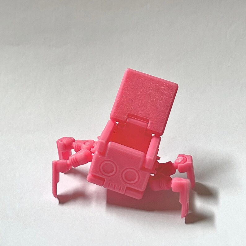 May include: A bright pink plastic articulated robot figure with a boxy head featuring two circular eyes and a rectangular mouth. The robot has a hinged top section that opens upwards and four jointed legs extending from the central body structure.