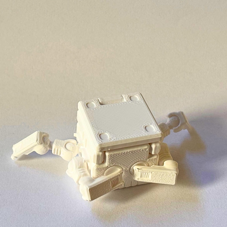 May include: A white 3D printed articulated cube figure with a square top surface featuring four circular indentations at the corners. The object has multiple jointed limbs extending from the sides, showcasing a layered texture from the printing process.