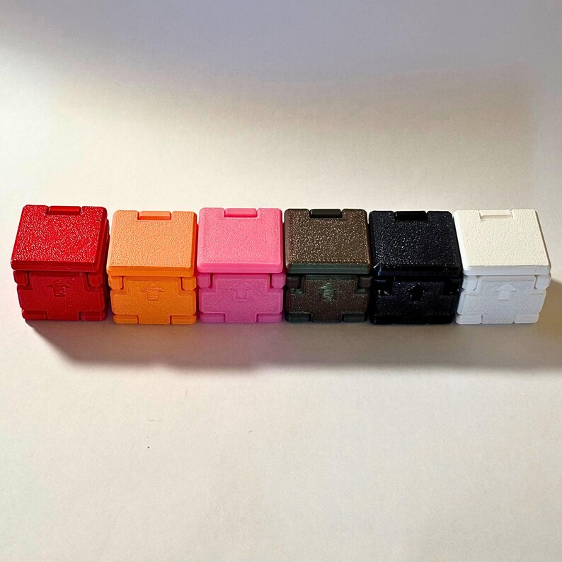 May include: A row of six small, square, hinged boxes in red, orange, pink, brown, black, and white. Each box features a textured surface and a small, moulded upward-pointing arrow symbol on the front panel, indicating the orientation for opening.