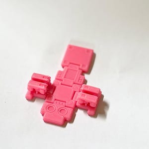 May include: A bright pink, articulated plastic figure with a blocky, geometric design. The object features a central torso with two circular indentations, two side appendages with mechanical details, and a rectangular top section.