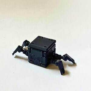 May include: A small, matte black, 3D-printed mechanical creature featuring a cubic body with a skull-like face design, two circular eye indentations, and four articulated, jointed legs extending from the sides of the central block structure.