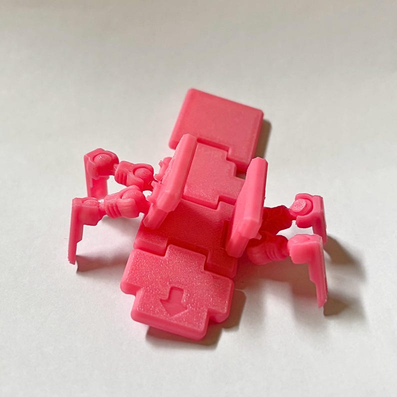 May include: A bright pink articulated plastic figure featuring a central rectangular body with a moulded arrow pointing downwards. The figure has four jointed, mechanical-style legs extending from the sides and a flat, rectangular head piece.