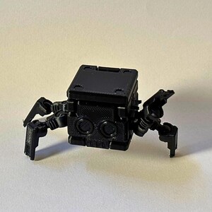 May include: A small, black, 3D-printed robotic figure with a cubic body and four articulated legs. The front of the cube features two circular eye-like indentations and a textured surface, while the top is flat with recessed corner details.