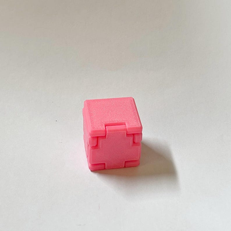 May include: A small, solid pink cube featuring a distinct cross-shaped indentation on its front face. The object has a matte texture and a geometric, blocky design with sharp, defined edges and corners, presented as a single, uniform unit.