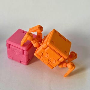 May include: A bright orange articulated robot figure with a square body and jointed limbs positioned next to a solid pink cube. The robot features circular details on its front panel, while the pink cube displays a moulded arrow symbol on its side.