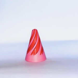 May include: A conical object featuring a smooth, light pink base and a series of raised, swirling red ridges that spiral upward toward the pointed top. The design creates a distinct texture with alternating colors and a symmetrical shape.