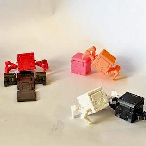 May include: A collection of six small, articulated cube-shaped robot figures in red, brown, pink, orange, white, and black. Each figure features a square head with two circular eyes and jointed limbs, displayed in various poses on a plain surface.