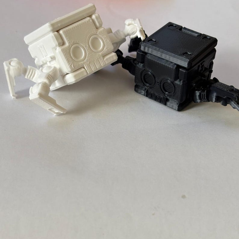 May include: Two small, articulated robot figurines, one in matte white and one in matte black. Each robot features a cube-shaped head with two circular eye-like indentations, a textured top surface, and jointed limbs extending from the sides.