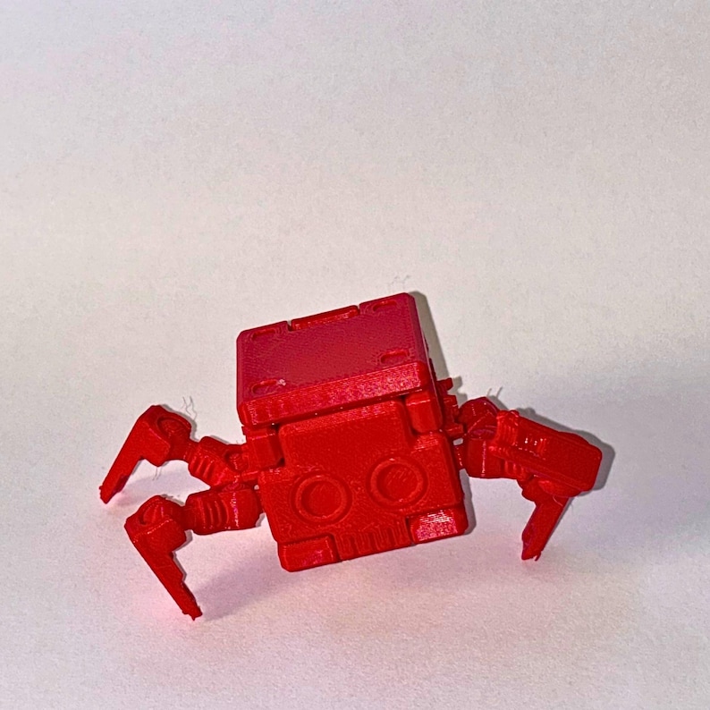 May include: A bright red, articulated robotic creature featuring a square-shaped body with two circular eye-like indentations on the front face. The figure has multiple jointed, mechanical-style legs extending from the sides of the central block.