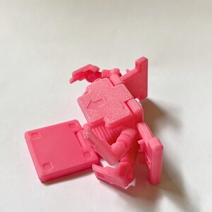May include: A bright pink plastic articulated robot figure featuring a blocky design with a flat base, jointed limbs, and a square torso section. The chest area displays a moulded arrow pointing upward, and the surface has a textured finish.