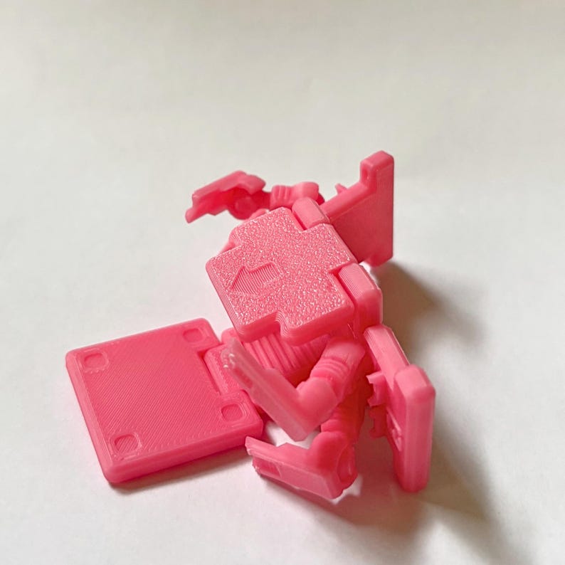 May include: A bright pink plastic articulated robot figure featuring a blocky design with a flat base, jointed limbs, and a square torso section. The chest area displays a moulded arrow pointing upward, and the surface has a textured finish.