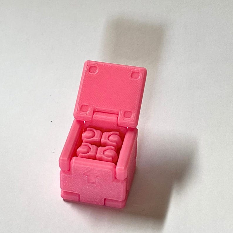 May include: A small, bright pink, 3D-printed plastic box with an open hinged lid. Inside the box, four small, identical, pink, articulated, humanoid-shaped figures are arranged in a square formation. The front of the box features an arrow.