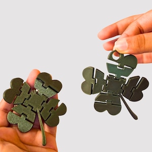 May include: Two dark olive green four-leaf clover shapes constructed from articulated, interlocking segments. Each clover features a central square piece with four radiating leaf sections connected by hinges, allowing for flexible movement and folding.