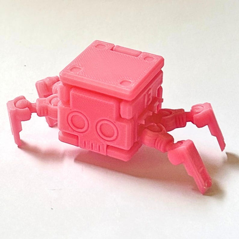 May include: A bright pink, small, articulated robot figurine with a square-shaped head and body. The robot features two circular eye-like indentations on the front face and four jointed, multi-segmented legs extending from the sides of the body.