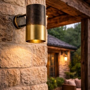 May include: A cylindrical outdoor wall sconce with a two-tone finish. The top portion is dark bronze, and the bottom is gold. The sconce is mounted on a stone wall with a warm, inviting glow. The fixture is approximately 10 inches tall.