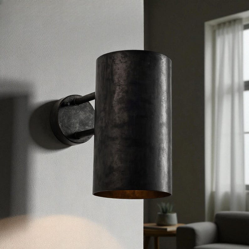May include: A cylindrical, dark metal wall sconce with a distressed finish. The sconce is mounted on a circular base and has a warm, golden interior. The design is industrial and minimalist, suitable for interior lighting.