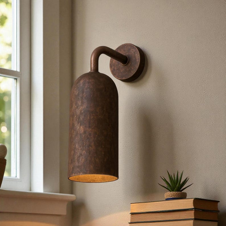 Pu&ograve; includere: Applique da parete color bronzo con paralume cilindrico e braccio curvo. L'applique &egrave; montata su una base circolare. Il paralume ha una superficie strutturata e un bagliore caldo. Sono visibili una piccola pianta e dei libri.