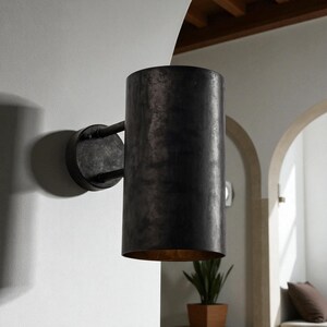 May include: A cylindrical, dark metal wall sconce with a distressed finish. The sconce has a circular base and a curved arm connecting to the cylindrical shade. The interior of the shade has a gold finish. The sconce is mounted on a white wall.
