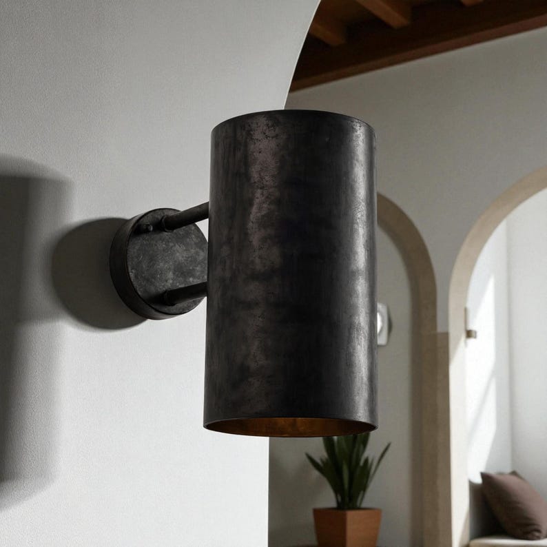 May include: A cylindrical, dark metal wall sconce with a distressed finish. The sconce has a circular base and a curved arm connecting to the cylindrical shade. The interior of the shade has a gold finish. The sconce is mounted on a white wall.