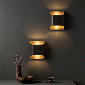 K&ouml;nnte beinhalten: Zwei moderne, rechteckige Wandleuchten mit schwarzem Finish und goldenen Akzenten, die ein warmes Licht ausstrahlen. Die Leuchten sind an einer dunkelgrauen Wand angebracht. Eine dekorative Vase, eine Schale und eine Skulptur befinden sich auf einer dunkelgrauen Oberfl&auml;che.