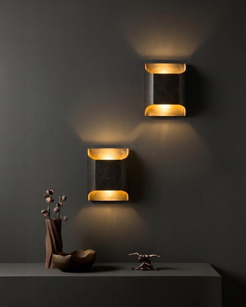 K&ouml;nnte beinhalten: Zwei moderne, rechteckige Wandleuchten mit schwarzem Finish und goldenen Akzenten, die ein warmes Licht ausstrahlen. Die Leuchten sind an einer dunkelgrauen Wand angebracht. Eine dekorative Vase, eine Schale und eine Skulptur befinden sich auf einer dunkelgrauen Oberfl&auml;che.