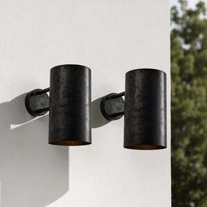 May include: Two cylindrical, dark bronze outdoor wall sconces. Each sconce has a circular mounting plate and a vertical, open-ended shade. The interior of the shades have a golden hue. The sconces are mounted on a white wall.