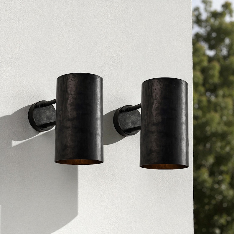 May include: Two cylindrical, dark bronze outdoor wall sconces. Each sconce has a circular mounting plate and a vertical, open-ended shade. The interior of the shades have a golden hue. The sconces are mounted on a white wall.