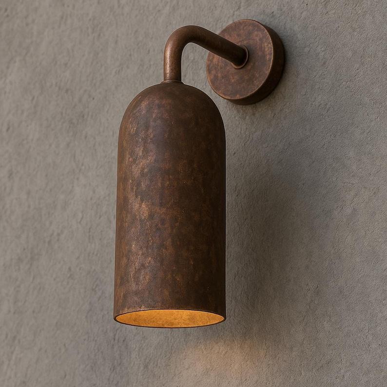 Pu&ograve; includere: Applique da parete in bronzo con paralume cilindrico e braccio curvo. L'applique ha una piastra posteriore circolare e una calda luce che emana dal basso. Montato su una parete grigia strutturata.