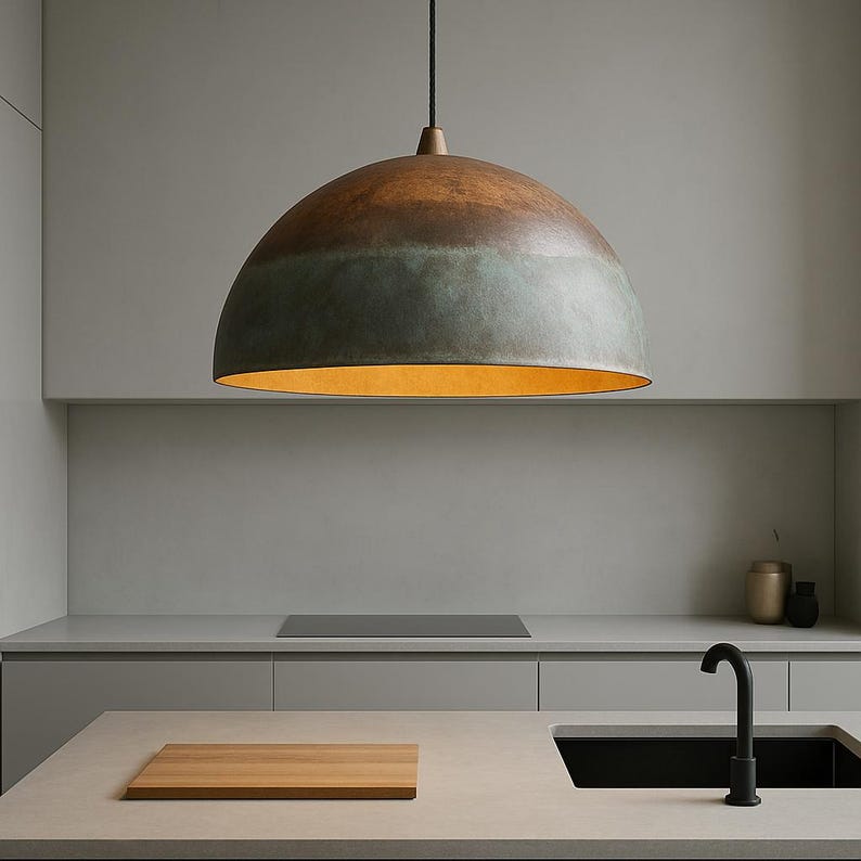 May include: A large, dome-shaped pendant light fixture with a two-tone finish of brown and green. The interior of the light is a warm yellow. The light hangs above a kitchen countertop with a wooden cutting board and a black faucet.