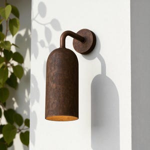 Pu&ograve; includere: Applique da parete per esterni color bronzo con paralume cilindrico e braccio curvo. Montato su un muro bianco, proietta un'ombra. L'apparecchio ha una finitura strutturata e un bagliore caldo.
