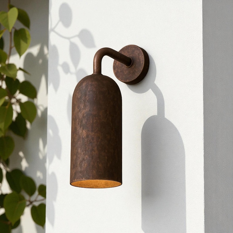 Pu&ograve; includere: Applique da parete per esterni color bronzo con paralume cilindrico e braccio curvo. Montato su un muro bianco, proietta un'ombra. L'apparecchio ha una finitura strutturata e un bagliore caldo.