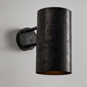 May include: A cylindrical, dark metal wall sconce with a distressed finish. The sconce has a circular base and a long, tubular shade. The interior of the shade has a warm, golden hue. The fixture is mounted on a white wall.