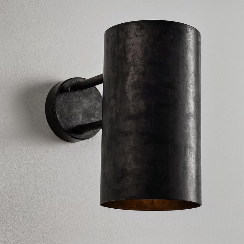 May include: A cylindrical, dark metal wall sconce with a distressed finish. The sconce has a circular base and a long, tubular shade. The interior of the shade has a warm, golden hue. The fixture is mounted on a white wall.