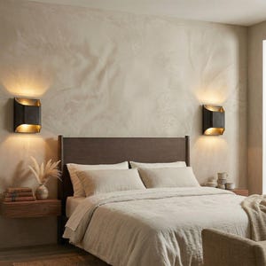 K&ouml;nnte beinhalten: Schlafzimmer-Interieur mit Bett, Nachttischen und Wandleuchten. Das Bett hat eine beige Bettdecke und Kissen. Zwei schwarze und goldene Wandleuchten sind an der Wand montiert. Ein Nachttisch aus Holz enth&auml;lt B&uuml;cher und eine Vase mit getrockneten Blumen.